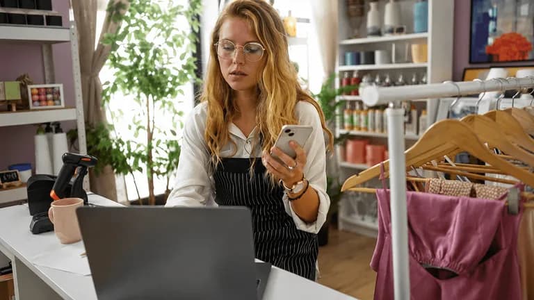 Microempreendedora de loja de roupas checando o celular em frente ao laptop enquanto atende o balcão da loja
