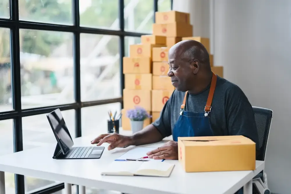 Microempreendedor analisando relatórios no tablet ao lado de caixas de pedidos.
