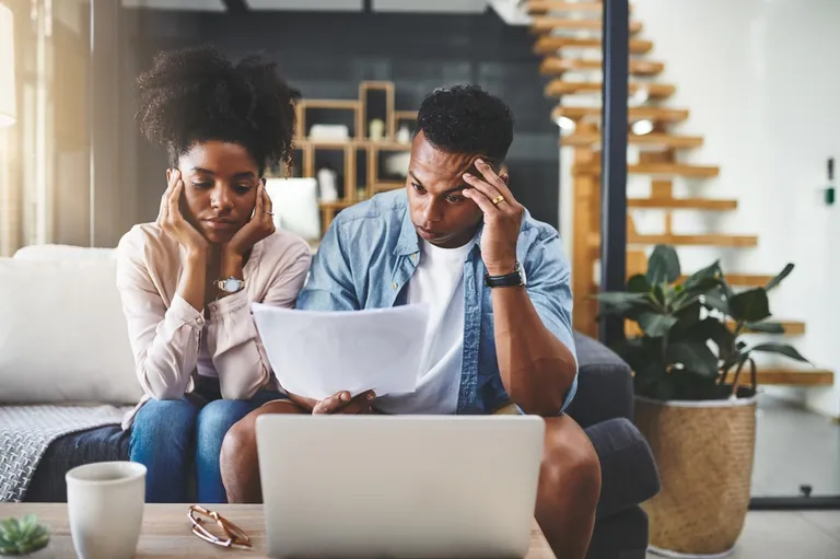 A imagem mostra um casal sentado em um sofá cinza, em um ambiente doméstico moderno e bem iluminado. Ambos parecem preocupados ou estressados: a mulher, com cabelo cacheado preso, apoia o rosto nas mãos enquanto olha para um papel; o homem, ao lado dela, segura a cabeça com uma das mãos e olha para uma folha de papel que está segurando, com uma expressão tensa, como quem está analisando contas ou boletos.  Na mesa de centro à frente deles há um notebook aberto, uma xícara branca e um óculos apoiado. Ao fundo, é possível ver uma escada de madeira, uma estante decorativa e um vaso com planta, compondo um cenário de sala de estar aconchegante.  A cena transmite a sensação de preocupação financeira, o tipo de imagem usada para ilustrar temas como organização de contas a pagar, planejamento financeiro ou dificuldades no controle do orçamento doméstico ou de um pequeno negócio.