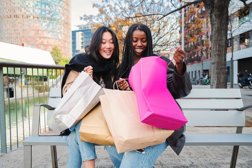 Duas jovens sorridentes sentadas em um banco na rua, segurando sacolas de compras coloridas durante as promoções do Dia do Cliente 2026