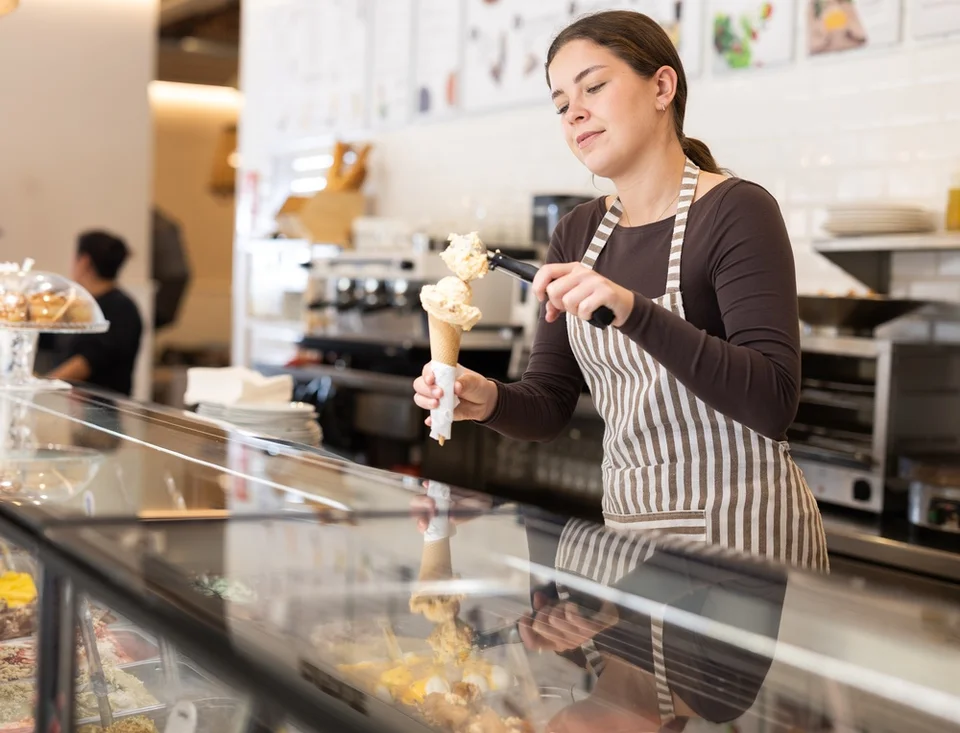 Atendente servindo sorvete de casquinha no balcão de uma sorveteria com vitrine de sabores