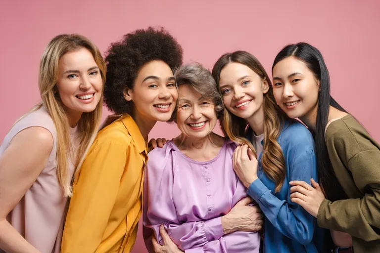 Cinco mulheres sorridentes de diferentes idades e etnias abraçadas em fundo rosa — Promoção no Dia das Mulheres
