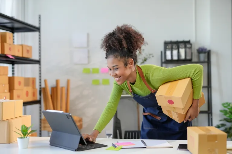 Empreendedora organizando caixas e atualizando o inventário de estoque em um tablet dentro de seu pequeno negócio.