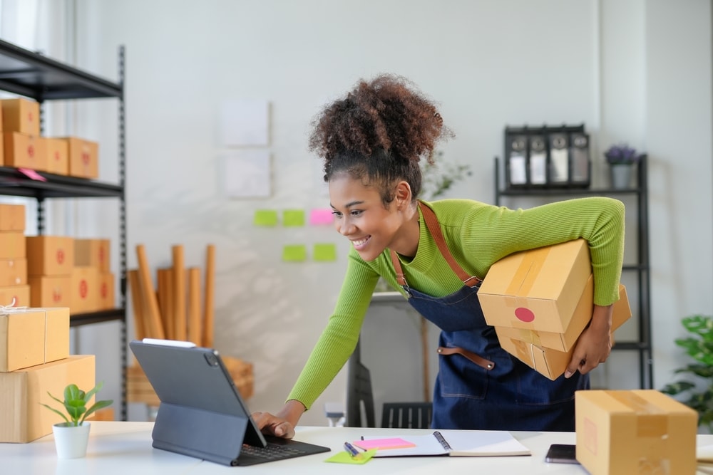 Empreendedora organizando caixas e atualizando o inventário de estoque em um tablet dentro de seu pequeno negócio.