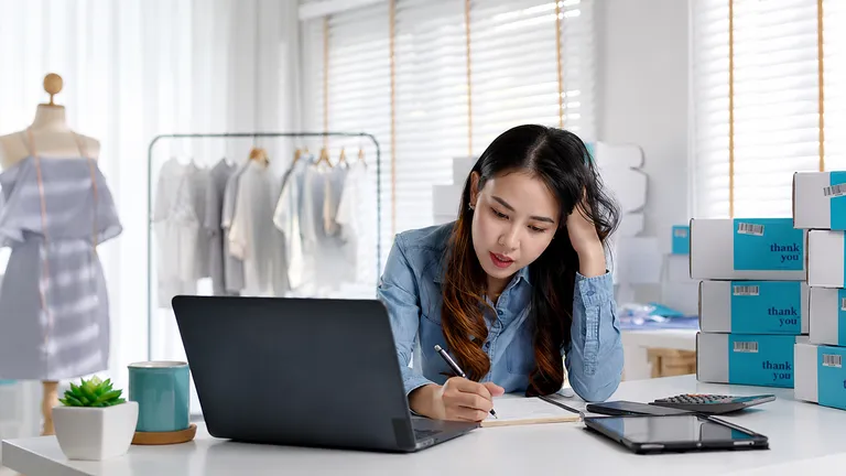 Microempreendedora de loja de roupas parece sobrecarregada enquanto anota contas à mão, com laptop, calculadora e caixas de produtos ao redor do espaço de trabalho.