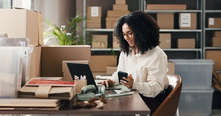 Empreendedora organizando pedidos no notebook e celular em estoque com caixas, representando como centralizar estoque, vendas e finanças em um só lugar.