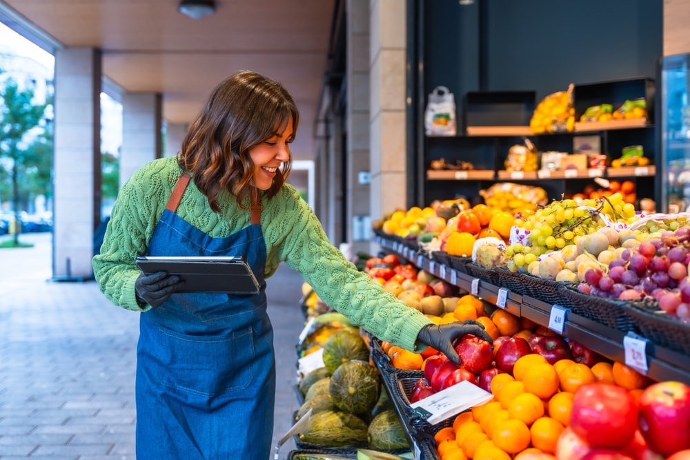 Empreendedora organizando produtos em prateleira de frutas enquanto confere estoque em tablet.